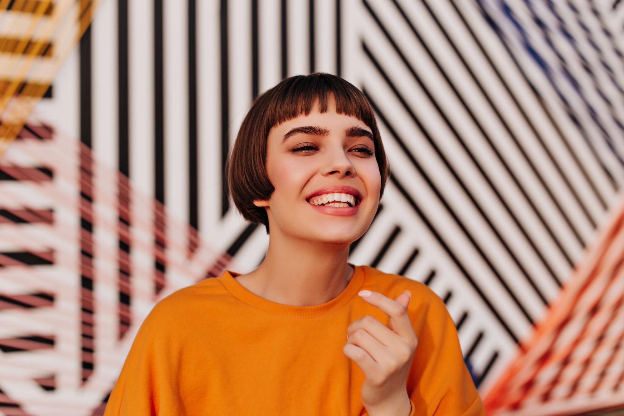 Optimistic teen girl in orange sweatshirt smiling on striped backdrop. Brunette woman in bright out