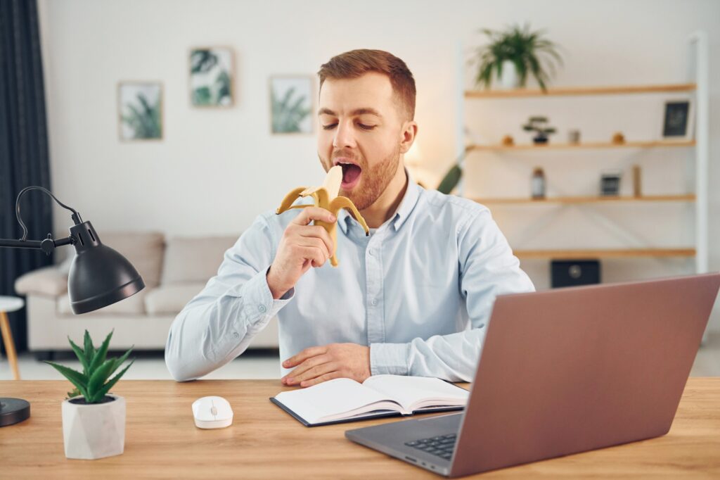 Eating the fruit. Man with banana sitting by the table with laptop on it and have fun