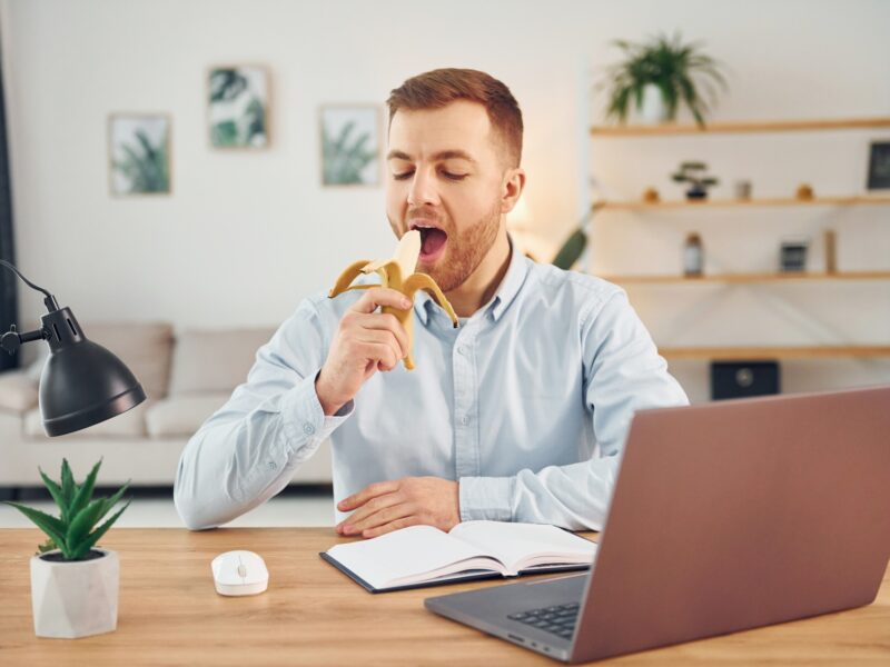 Eating the fruit. Man with banana sitting by the table with laptop on it and have fun